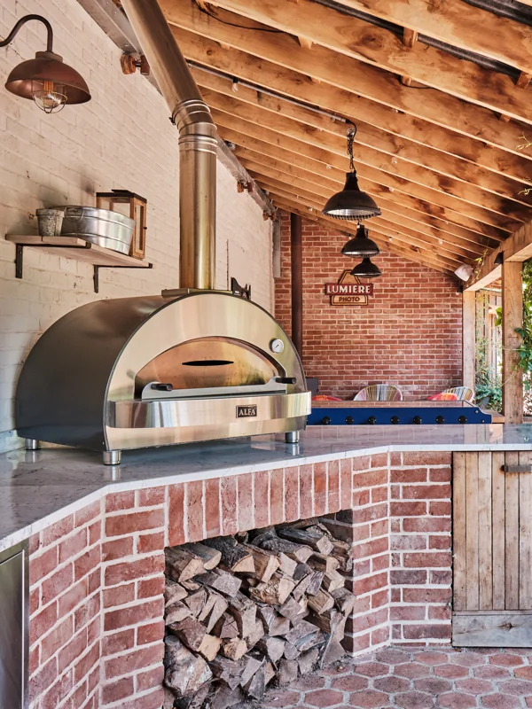 Brick outdoor kitchen with pizza oven, stacked logs and a timber roof